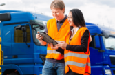 a man and woman, both wearing high-vis, stand in front of a line of blue HGVs consulting a tablet device.