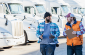 two men walk in front of a fleet of HGV vehicles. One wears a cap and orange high vis. he carries a clipboard.