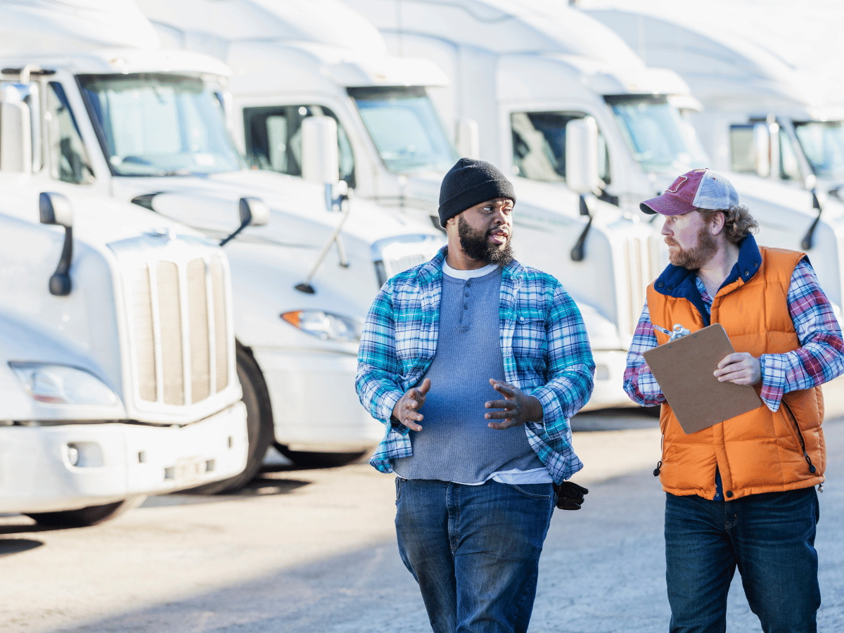 two men walk in front of a fleet of HGV vehicles. One wears a cap and orange high vis. he carries a clipboard.