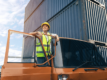 a female hgv driver stands leaning our of her cab with the door open. She wears a high vis vest and hard hat.