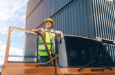 a female hgv driver stands leaning our of her cab with the door open. She wears a high vis vest and hard hat.