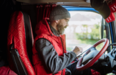 A male HGV driver wearing a woolly hat and red gillet sits in the drivers seat. He is filling out a test form on a device.