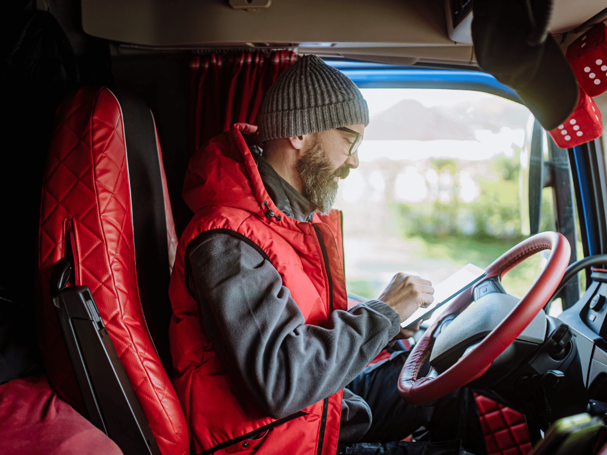 A male HGV driver wearing a woolly hat and red gillet sits in the drivers seat. He is filling out a test form on a device.